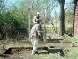 The archaeologist tosses soil from the test unit into the sifting screen hanging from the frame. The soil is then passed through the wire mesh of the screen for easy recovery of artifacts.