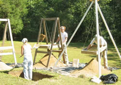 Soil from the test unit is heaved into the sifting screen while the archaeologist on the right searches through her screen for artifacts.